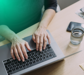 Hands typing on laptop at desk with notebook, phone, and water glass.