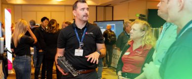 Two men shaking hands at networking event with booths and attendees in background.