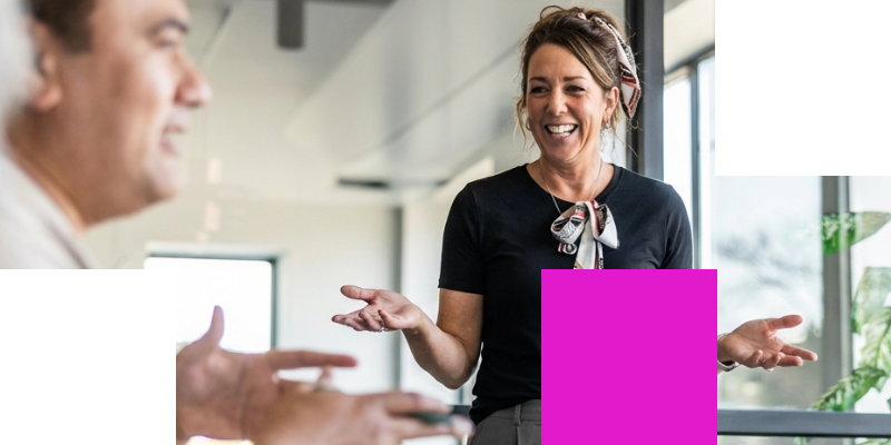Smiling woman gesturing while speaking with a colleague in a bright, modern office.