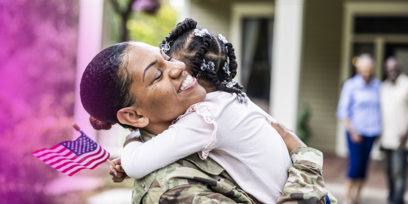 A military service member in uniform hugging a young child outside a home, with two adults in the background.