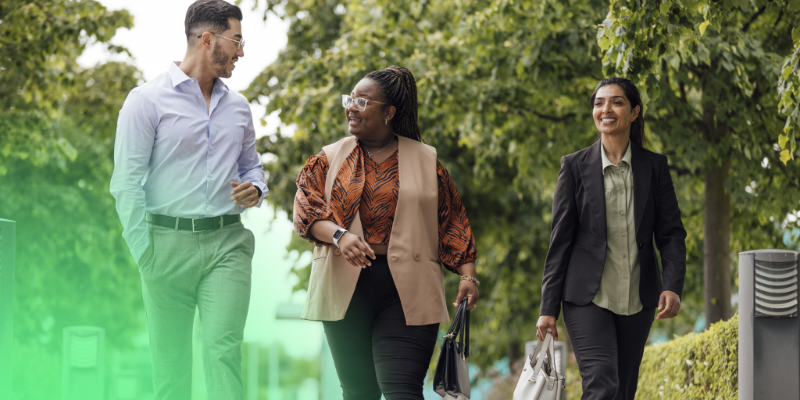 Three colleagues walking outdoors together, talking and smiling in a green park setting.