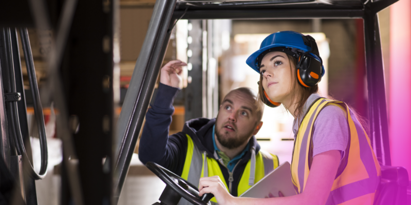 A young worker in safety gear (hard hat and reflective vest) smiling while talking with a coworker on a bus or work vehicle.