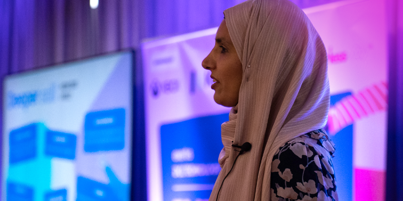 A woman wearing a headscarf speaking at an event, with a presentation screen blurred in the background.