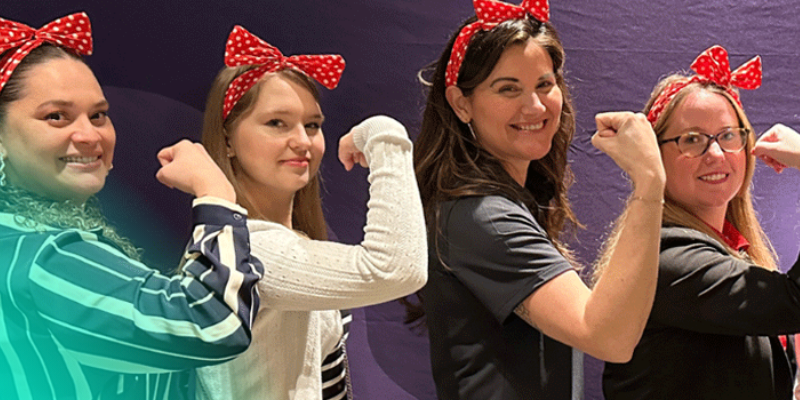 Four women standing together wearing red bandanas, smiling and flexing their arms in a gesture of empowerment.