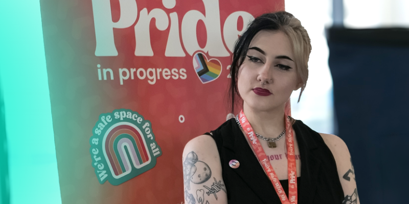 A person with short hair standing near a sign that reads “Pride in progress,” looking toward the camera.