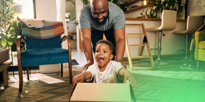 A father smiling and playing with a young child who is crawling on the floor in a home setting.