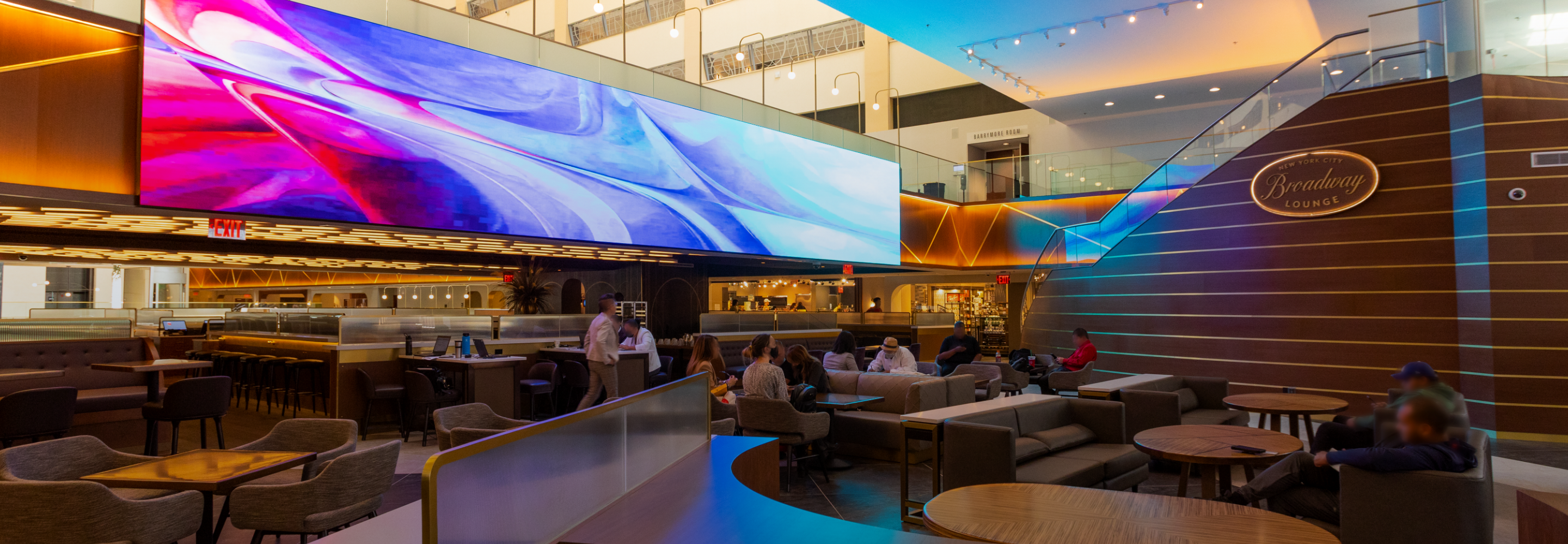 Broadway Lounge interior with modern seating, large digital screen, and people conversing.