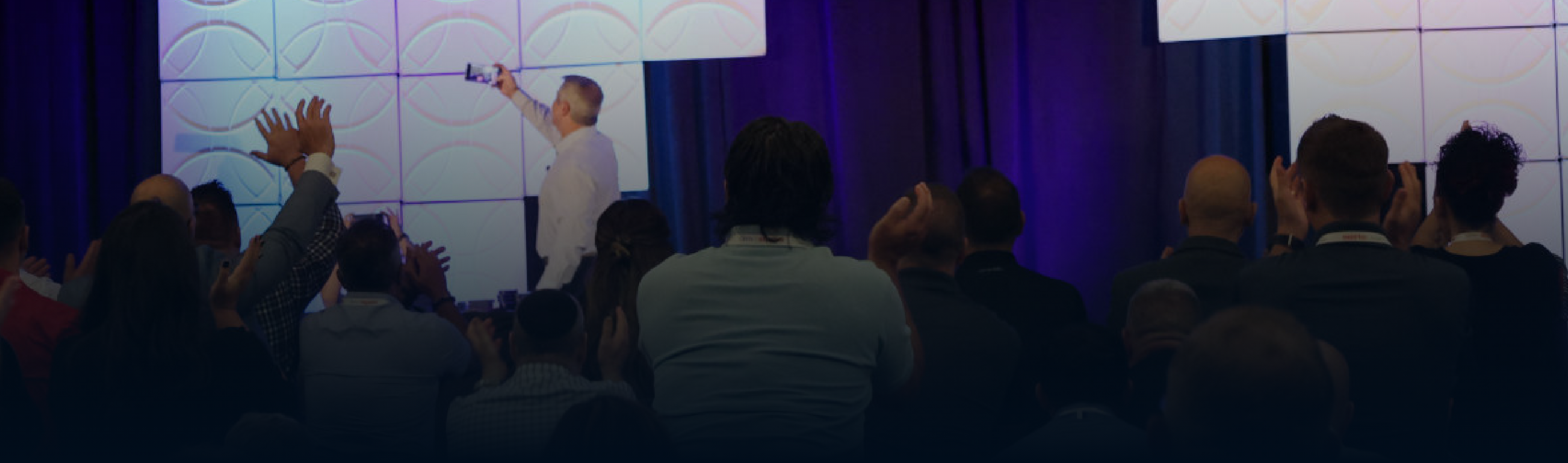 Man presenting on stage with pastel curved-line display, audience raising hands.