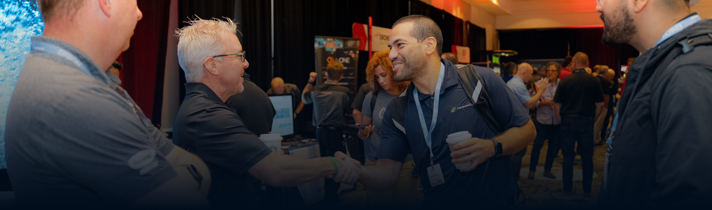 Two men shaking hands at networking event with booths and attendees in background.
