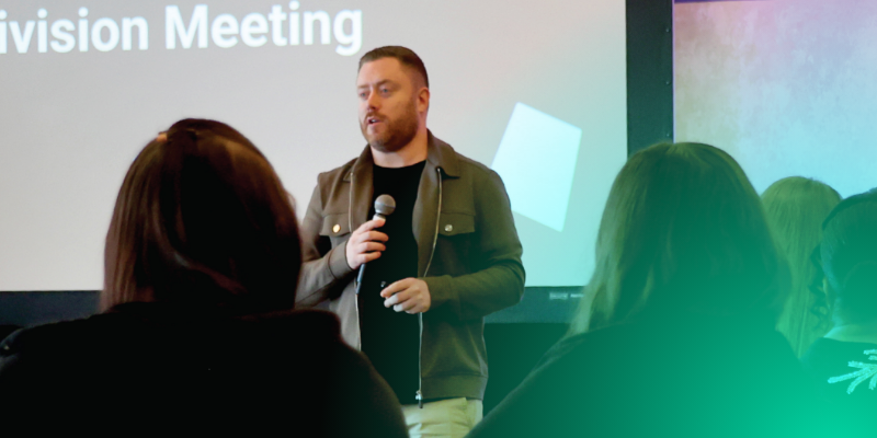 Man speaking at Division Meeting with audience and screen behind.