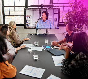 Team in conference room watching a woman speak on a large video screen.